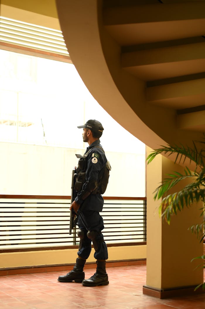 A security guard in uniform stands indoors, observing through a window by a staircase.
